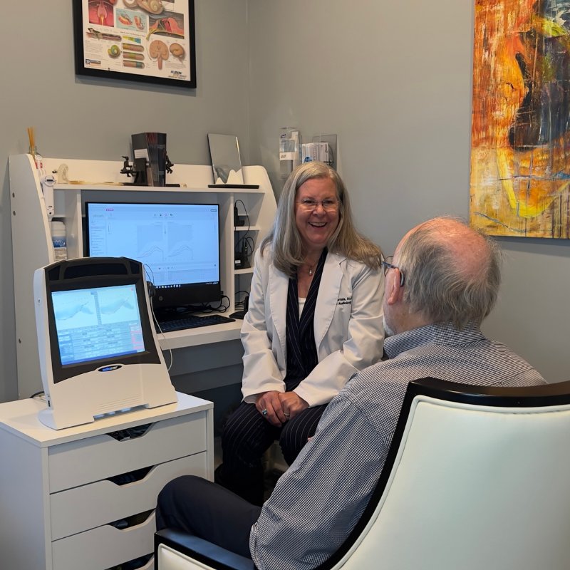 Dr. Thomas, in a white coat, smiling as she talks with a patient beside her hearing test equipment