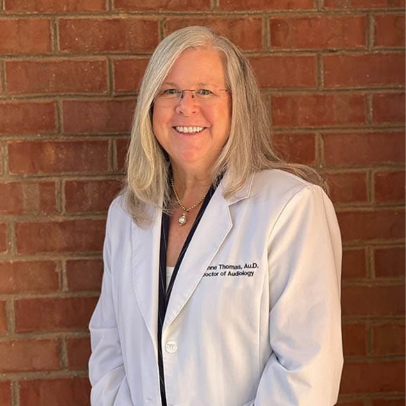 Dr. G'Anne Thomas, audiologist, smiling in glasses and a white Doctor of Audiology coat in front of a brick wall